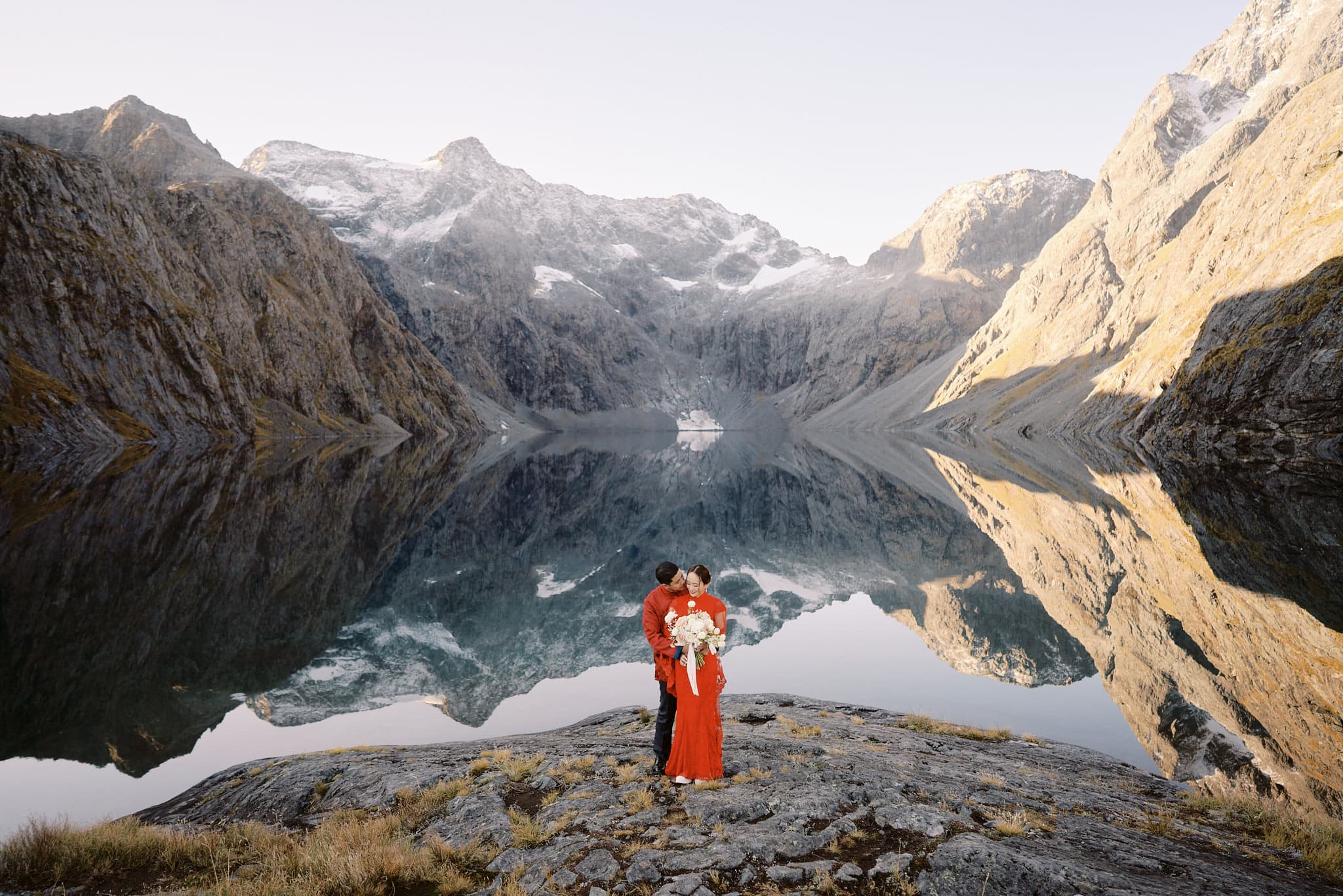 クイーンズタウン　ニュージーランド　ウェディング　前撮り　挙式　結婚式　フォトグラファー | ニュージーランドの湖の前に立つ新婦と新郎付きのクイーンズタウン・ヘリウェディング＆エロープメントパッケージ。